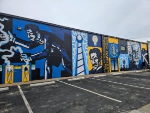 A Building Mural in Hays, KS showcases blue and yellow illustrations of scientists like Nikola Tesla and Albert Einstein, along with a light bulb, promoting STEM Harvest, with parking spaces in front and a cloudy sky above.