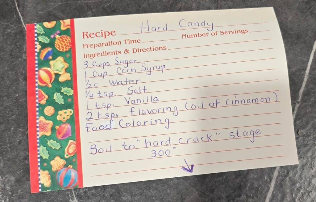 A handwritten recipe card for Linda's Hard Candy rests on a dark surface. Ingredients include sugar, corn syrup, water, salt, vanilla, flavoring, and food coloring. Instructions note to boil the mixture to 300°F (“hard crack” stage).