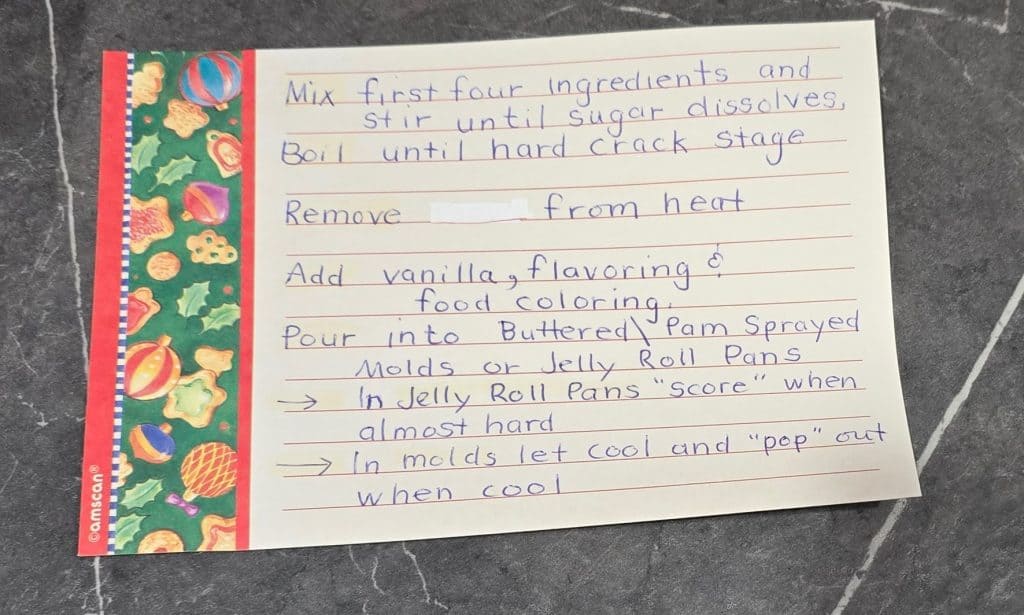 A handwritten recipe card on a dark countertop with a holiday-themed border lists steps for making Linda's Candy, including mixing sugar, boiling, adding flavor and color, and pouring the gourmet candy mixture into pans.