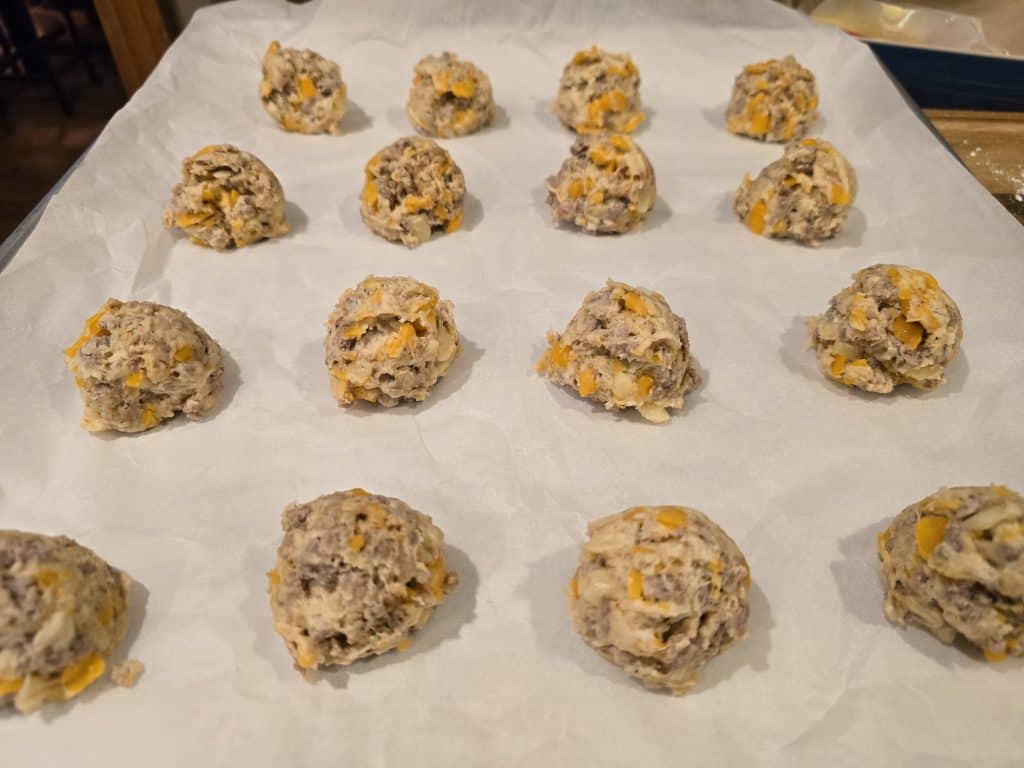 Scoops of unbaked cookie dough with visible chocolate chunks and nuts are arranged in rows on a parchment-lined baking sheet, just like Grandma's Sausage Bites lined up for a classic sausage appetizer.