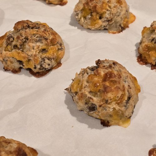 Drop biscuits with visible bits of sausage and melted cheese are arranged on parchment paper, freshly baked with golden-brown tops and crispy edges—just like Grandma's Sausage Bites, a classic sausage appetizer everyone will love.