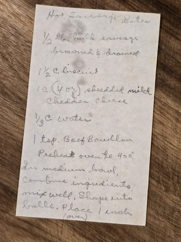 A handwritten Grandma's Sausage Bites recipe rests on white paper atop a wooden surface, detailing sausage, biscuit, cheese, water, and beef bouillon with instructions for mixing and baking this classic sausage appetizer.