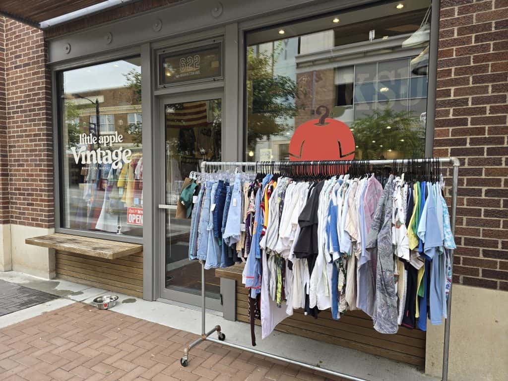 A clothing rack with assorted vintage clothes stands outside Little Apple Vintage Shop in Manhattan KS. Large windows display more items inside, and a red apple sign hangs above the entrance to this charming vintage shop.