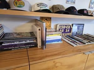 A wooden cabinet at Little Apple Vintage Shop in Manhattan KS displays hats, books, CDs, and DVDs. A sign reading "KSU-KU 48-16 10-18-1997" and various memorabilia are arranged neatly on top of the cabinet.