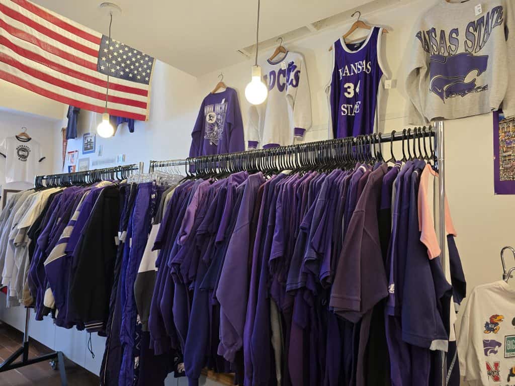 A clothing rack at Little Apple Vintage Shop in Manhattan KS is filled with mostly purple shirts and Kansas State University apparel. An American flag decorates the wall, while round lights hang from the ceiling, creating a cozy vintage shop vibe.