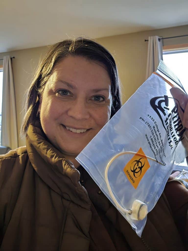 A smiling person in a brown jacket holds up a clear plastic biohazard bag containing a white medical device, capturing the moment of port removal—a milestone of both fear and relief—on their cancer journey, standing indoors with light curtains behind them.