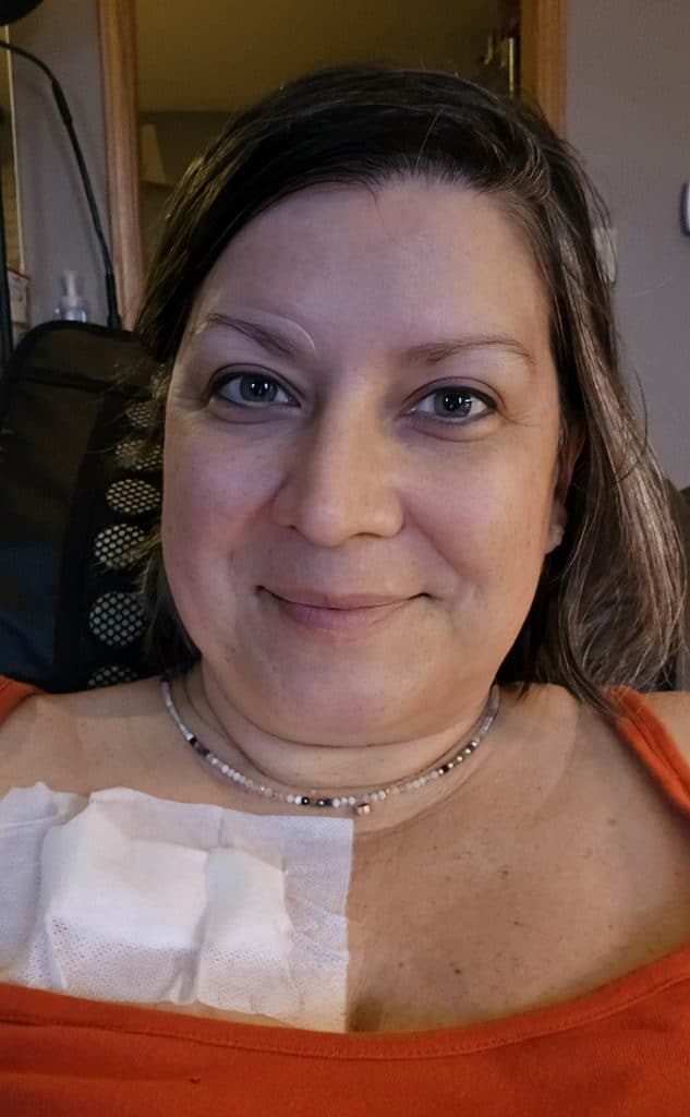 A woman with straight brown hair smiles softly at the camera. She is wearing an orange top and a beaded necklace. A white medical bandage on her upper chest hints at recent port removal during her cancer journey. The background shows a dimly lit room.