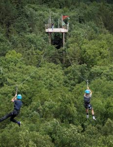 Two people wearing helmets and harnesses zipline over a dense green forest toward a wooden platform, where another person waits under a small roof and a red flag.