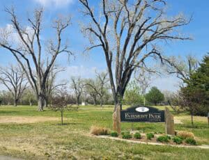 A park scene with leafless and budding trees, green grass, and a sign reading "Fairmont Park, 1701 South Plum St." under a blue sky with some clouds.
