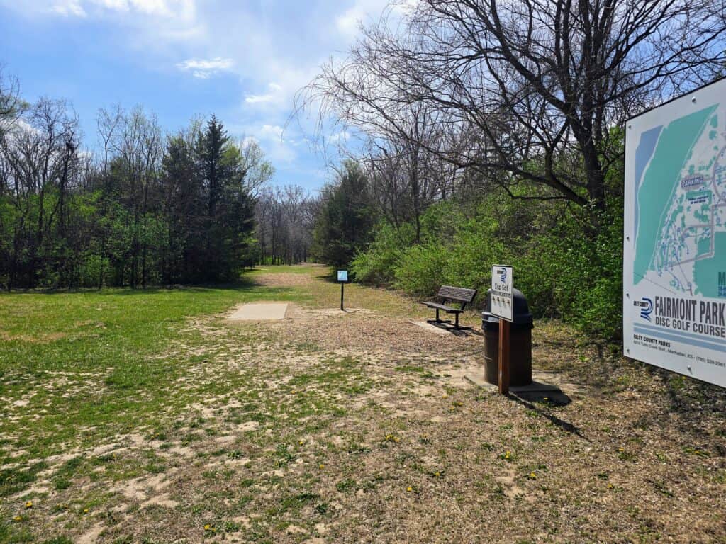 A disc golf course with a concrete tee pad, benches, informational signs, trees, and a large map on the right. The area is grassy with patches of dirt and surrounded by leafing trees under a partly cloudy sky.