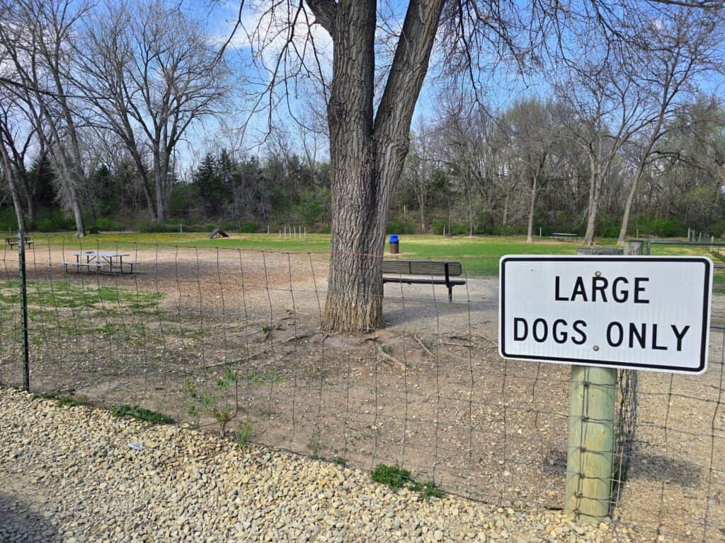 A fenced dog park area at Fairmont Park in Manhattan, Kansas, features a sign reading "Large Dogs Only." Inside the enclosure are benches, picnic tables, trees, and a trash can on mostly dirt with patches of grass.