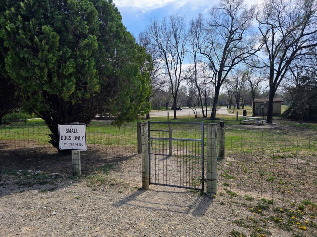 A fenced area with a gate in Fairmont Park, Manhattan Kansas, features a sign that reads "SMALL DOGS ONLY Less than 18 lbs." Trees and picnic tables can be seen in the background under a partly cloudy sky.