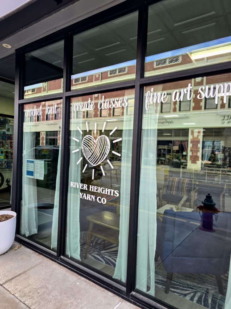 Storefront window of River Heights Yarn Co. in downtown Manhattan KS, featuring a heart logo and text promoting crochet, knit, create classes, fine art supplies, and a welcoming community; reflections of buildings and street scene are visible in the glass.