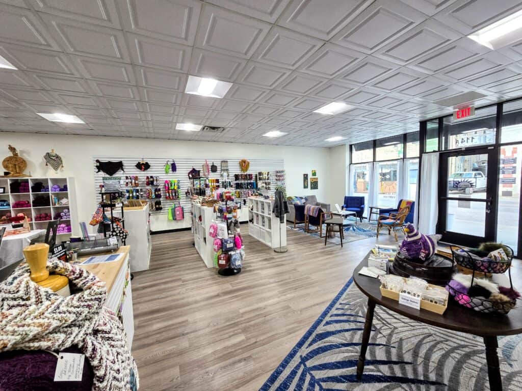 A bright, modern yarn shop in downtown Manhattan KS with shelves and tables displaying colorful yarn, knitting supplies, and handmade items. Large windows fill the cozy seating area near the entrance with natural light, perfect for community gatherings.
