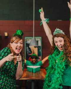 Two women in green outfits and festive accessories smile and celebrate St. Patrick&rsquo;s Day in Manhattan KS, as a man sits at a table with a beer pong setup; all appear to be enjoying the festivities together.