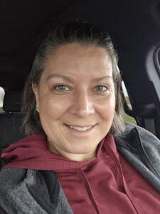 A cancer survivor with short brown hair and blue eyes, wearing a maroon hoodie and gray jacket, smiles while sitting in a car. The background shows the car&rsquo;s interior and a glimpse of life after chemo through the window.