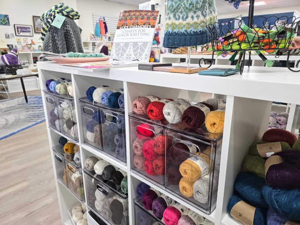 A white shelving unit holds clear bins filled with colorful yarn balls. Knitted hats and books are displayed on top. The background shows more yarn and knitting supplies in this bright downtown Manhattan KS craft store community.