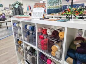 A white shelving unit holds clear bins filled with colorful yarn balls. Knitted hats and books are displayed on top. The background shows more yarn and knitting supplies in this bright downtown Manhattan KS craft store community.