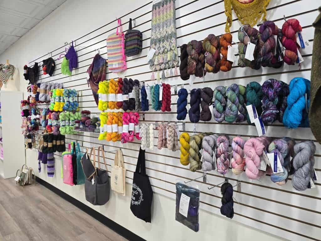 Wall display of colorful yarn skeins, knit samples, and tote bags in a modern Downtown Manhattan KS yarn shop. Yarns are arranged by color, with accessories and finished projects hanging above and below to inspire community creativity.