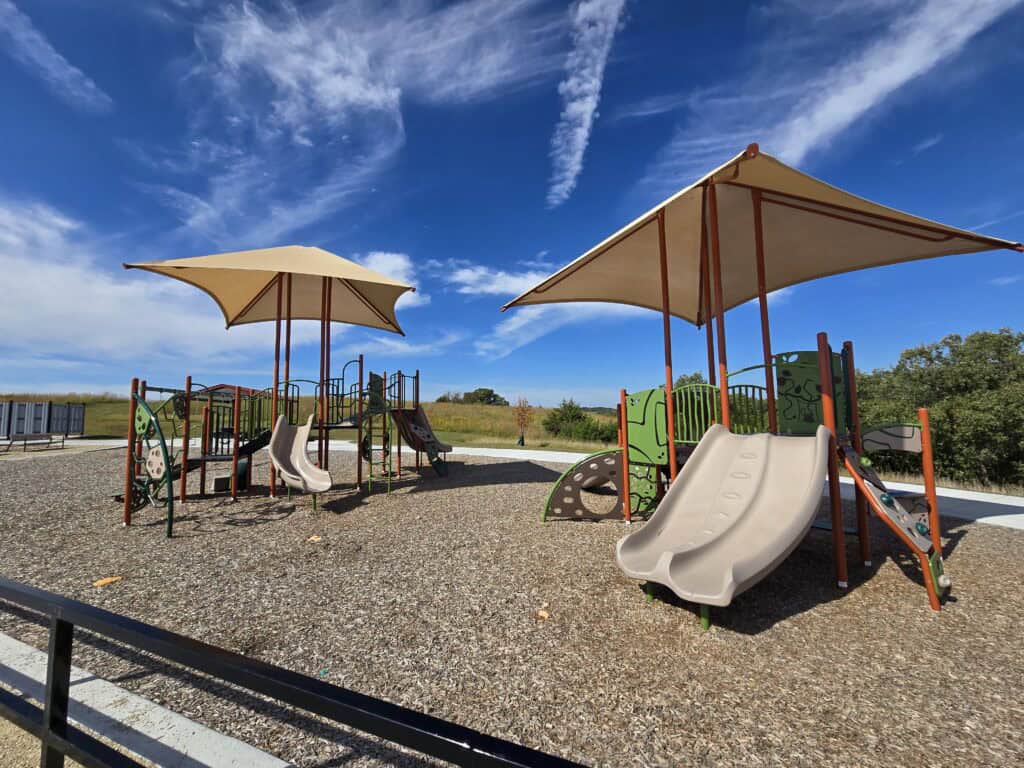 Set in Roger Schultz Community Park, this inviting playground in Manhattan KS features two large shade structures, slides, climbing equipment, and wood chip ground covering, all beneath a blue sky with wispy clouds, trees, and grassy areas nearby.