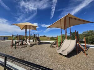 Set in Roger Schultz Community Park, this inviting playground in Manhattan KS features two large shade structures, slides, climbing equipment, and wood chip ground covering, all beneath a blue sky with wispy clouds, trees, and grassy areas nearby.