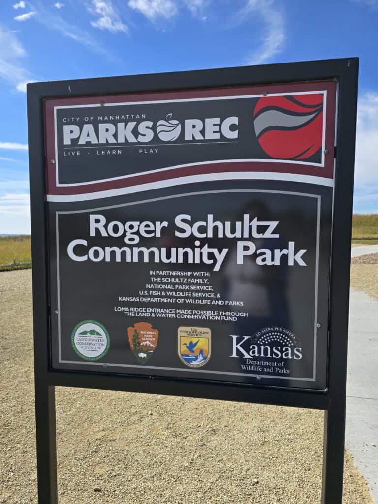A black and white outdoor sign reads “Roger Schultz Community Park” with the City of Manhattan KS Parks and Rec logo, partnership credits, and Kansas Department of Wildlife and Parks logos. The sign stands on a gravel path under a blue sky.