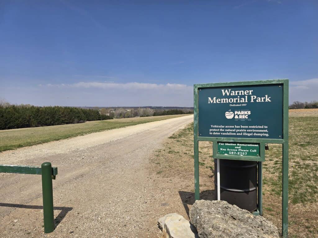 A green Warner Park sign stands beside a gravel road under a blue sky, with grassy fields and trees in the background. Located in Manhattan KS, it offers scenic Flint Hills views. A trash can sits next to the sign.