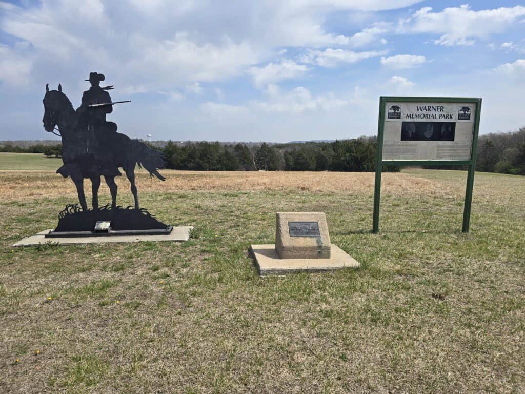 A metal silhouette of a horse and rider stands next to a plaque and a sign reading "Warner Memorial Park" in an open grassy field of the Flint Hills near Manhattan KS, under a partly cloudy sky.