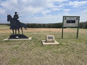 A metal silhouette of a horse and rider stands next to a plaque and a sign reading "Warner Memorial Park" in an open grassy field of the Flint Hills near Manhattan KS, under a partly cloudy sky.