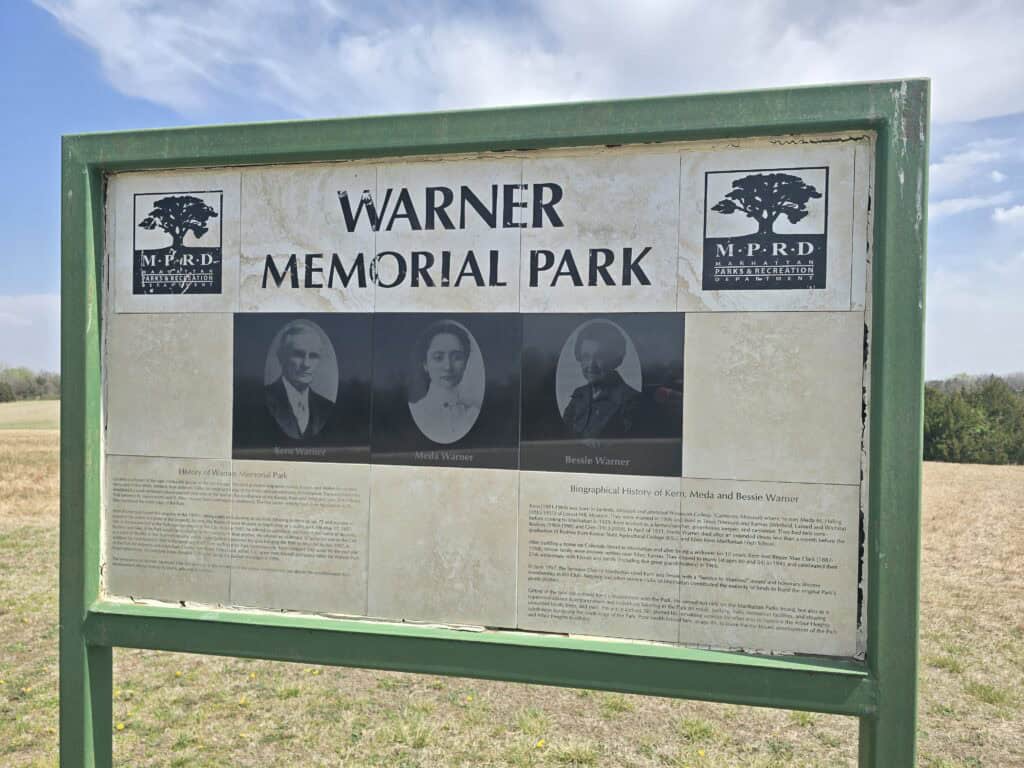 A weathered informational sign for Warner Park in Manhattan KS features black-and-white portraits of Bert, Mabel, and Bessie Warner—with historical text below and logos in the top corners, highlighting the park's ties to the Flint Hills.