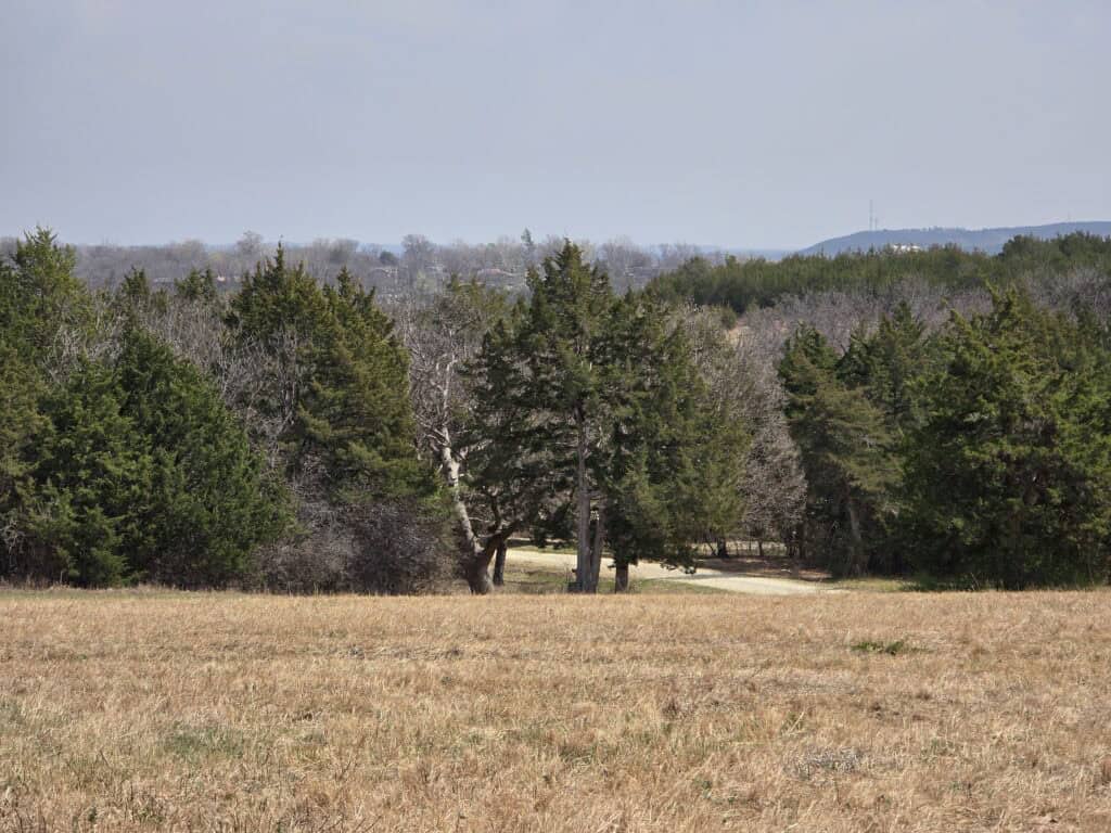 A dry, grassy field stretches into the distance at Warner Park, bordered by a line of green and leafless trees under a hazy sky. Low hills, part of the Flint Hills near Manhattan KS, are faintly visible in the background beyond the trees.