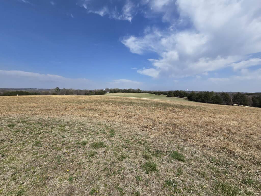 A wide, open field with dry grass and patches of green under a blue sky with scattered clouds, reminiscent of the Flint Hills near Manhattan KS. Sparse trees line the horizon in the distance.