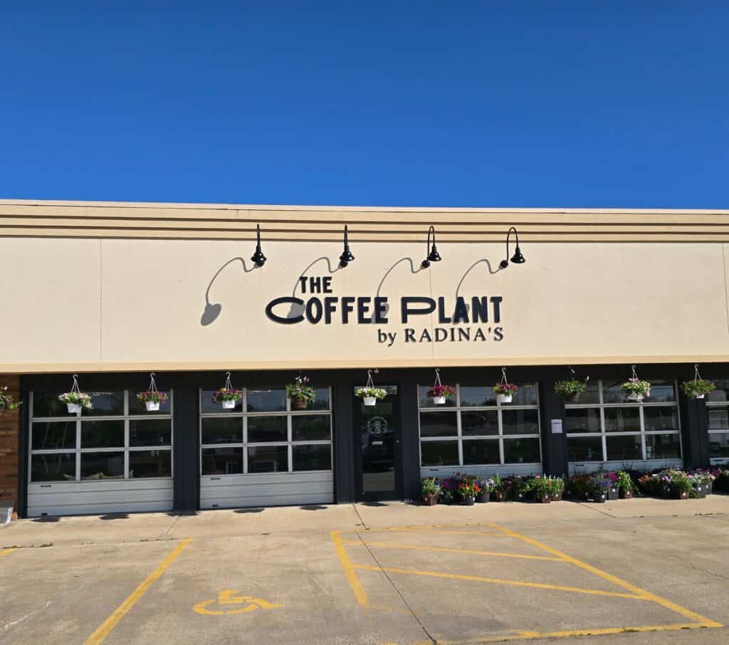 Exterior view of The Coffee Plant by Radina’s in Manhattan KS, featuring a beige facade, black sign, hanging lights, flower baskets, large windows, and a parking lot with a visible accessible space.