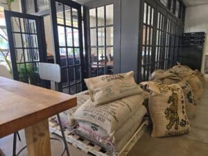 Large burlap sacks of Coffee Plant beans are stacked on wooden pallets inside Radina&rsquo;s caf&eacute; in Manhattan KS, next to black-framed glass doors and a wooden table. Natural light fills the cozy, industrial-style space.