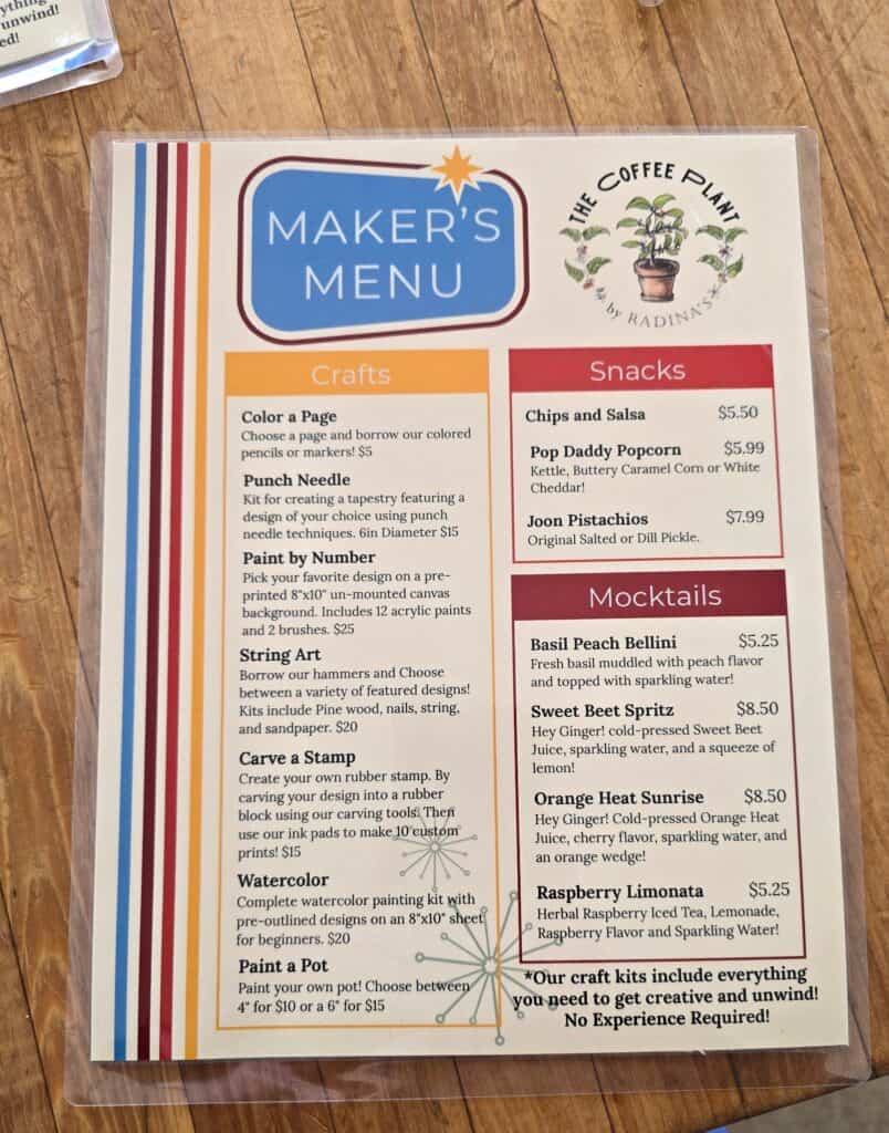 A colorful café menu at Radina’s in Manhattan KS sits on a wood table, listing craft activities, snacks, and mocktails. The crafts section features options like coloring, punch needle, and watercolor; the right side highlights snacks and drinks.