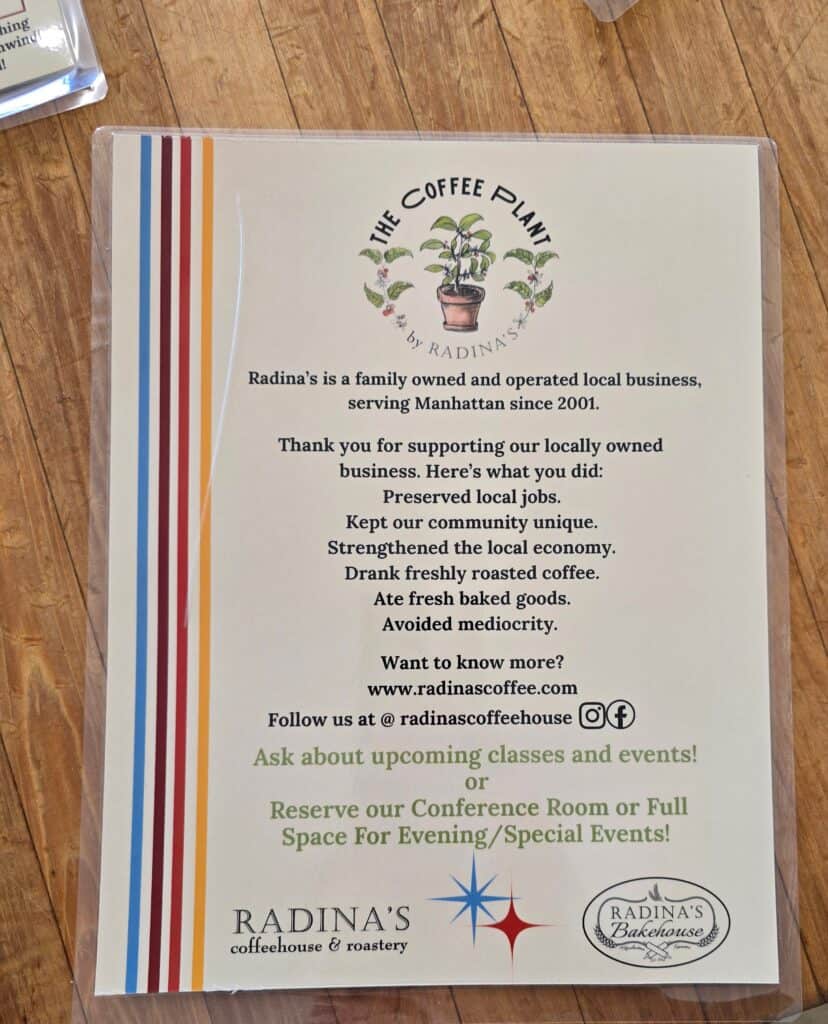 A flyer on a wooden table promotes Radina’s Coffeehouse in Manhattan KS, highlighting its local, family-owned roots since 2001, benefits of buying local from The Coffee Plant, event offerings, and social media details.