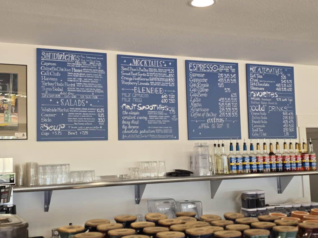 Three blue menu boards above the Radina’s café counter in Manhattan KS list sandwiches, salads, mocktails, espresso drinks made from Coffee Plant beans, smoothies, and hot alternatives. Below are glassware, coffee machines, bottles of syrup, and stacked mugs.