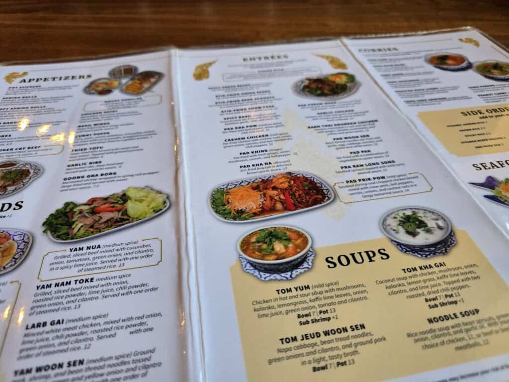 A close-up of the Baan Thai menu, one of the beloved Manhattan Kansas restaurants, displayed on a wooden table. The menu features appetizers, salads, curries, seafood, and more, with vibrant dish photos beside each description.