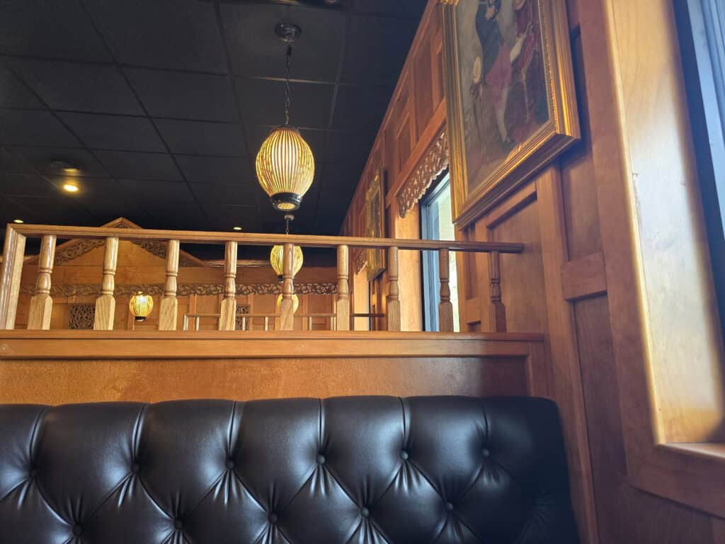 A cozy Thai restaurant interior with dark tufted leather seating, wooden paneling, hanging lantern-style lights, and framed artwork on the wall near a window letting in daylight—discover Baan Thai among Manhattan Kansas restaurants.