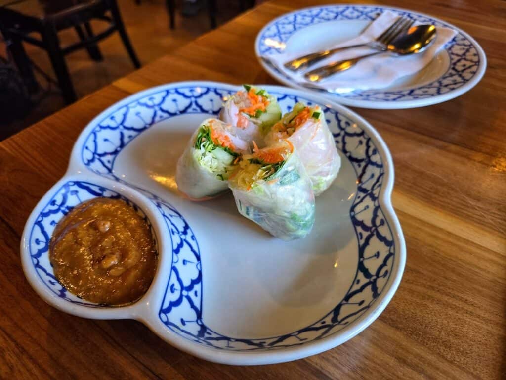 Three fresh spring rolls filled with vegetables are served on a patterned plate with peanut dipping sauce—a delicious appetizer from Baan Thai, one of the beloved Manhattan Kansas restaurants. In the background, another plate holds a fork and spoon on a wooden table.
