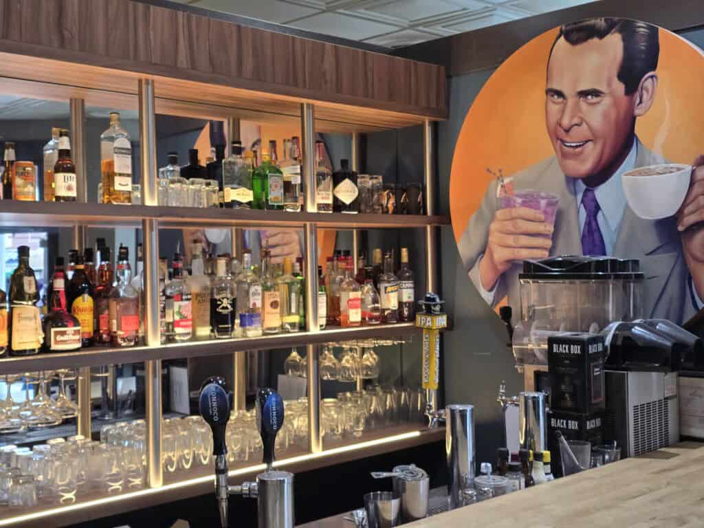 A bar with shelves of liquor bottles and glasses, beer taps, and cocktail shakers. On the wall is a large mural of a smiling man holding a cocktail and coffee—a nod to Manhattan KS and the vibrant spirit of Aggieville coffee shops like Bluestem Bistro.
