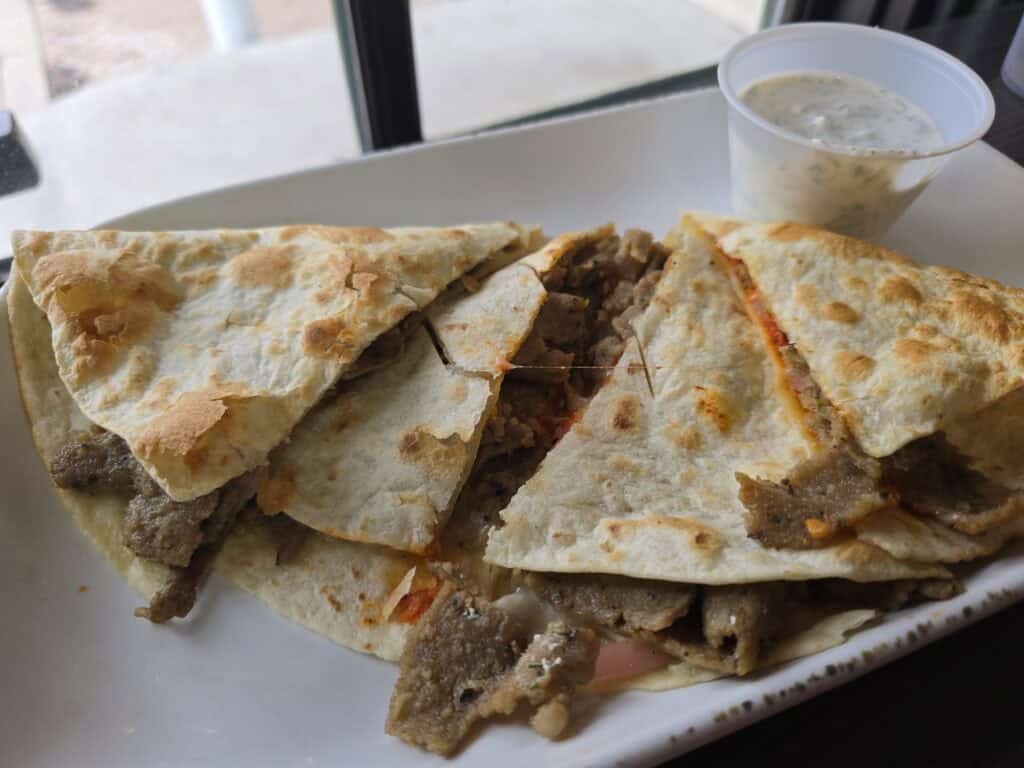 Two quesadilla wedges filled with seasoned meat are served on a white plate with a side of creamy dipping sauce in a small plastic cup. Enjoy this tasty dish near Manhattan KS coffee spots like Bluestem Bistro or while exploring Aggieville coffee shops.