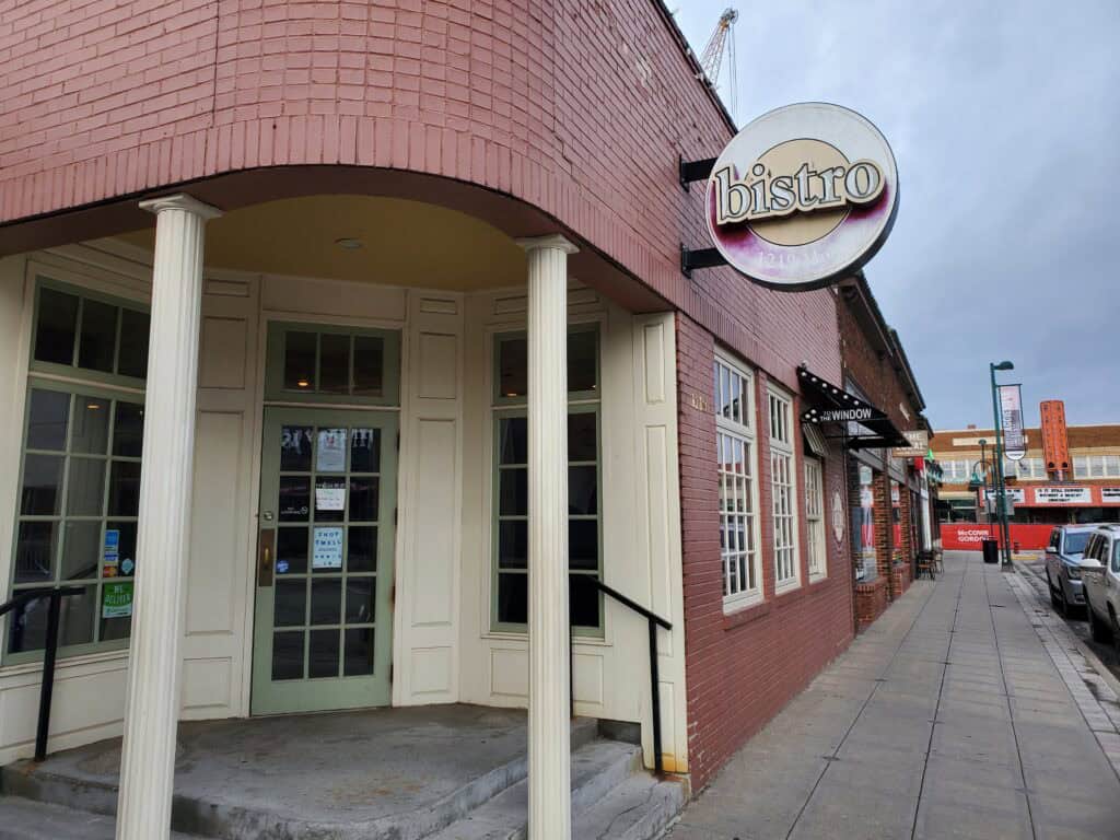 A brick building with cream-colored columns and a round sign reading “bistro” above the entrance. Located in Manhattan KS’s Aggieville, Bluestem Bistro stands out among local coffee shops, with a sidewalk running beside it and other buildings down the street.