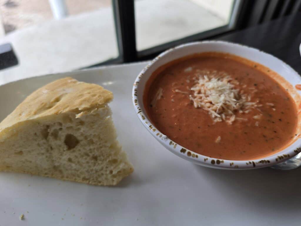 A bowl of creamy tomato soup topped with shredded cheese sits on a plate next to fresh bread at Bluestem Bistro, an inviting Aggieville coffee shop in Manhattan, KS. The setting is a cozy dining area near a sunlit window.